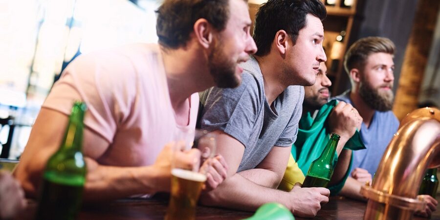 Vriendengroep kijkt gespannen naar een scherm tijdens een quiz met quotes in een café, met bierflessen op de bar